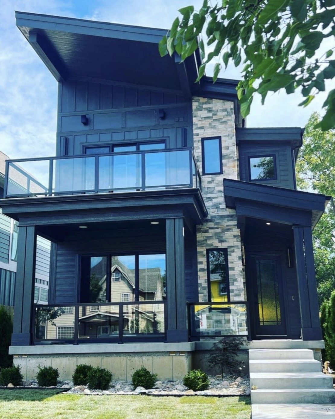 Modern three story custom home exterior in dark siding with stacked stone accents and a glass balcony rail