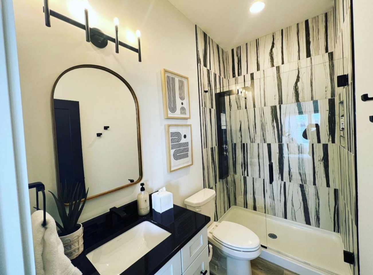 Black and white guest bath with dramatic veined marble shower surround, arched brass mirror and black quartz vanity