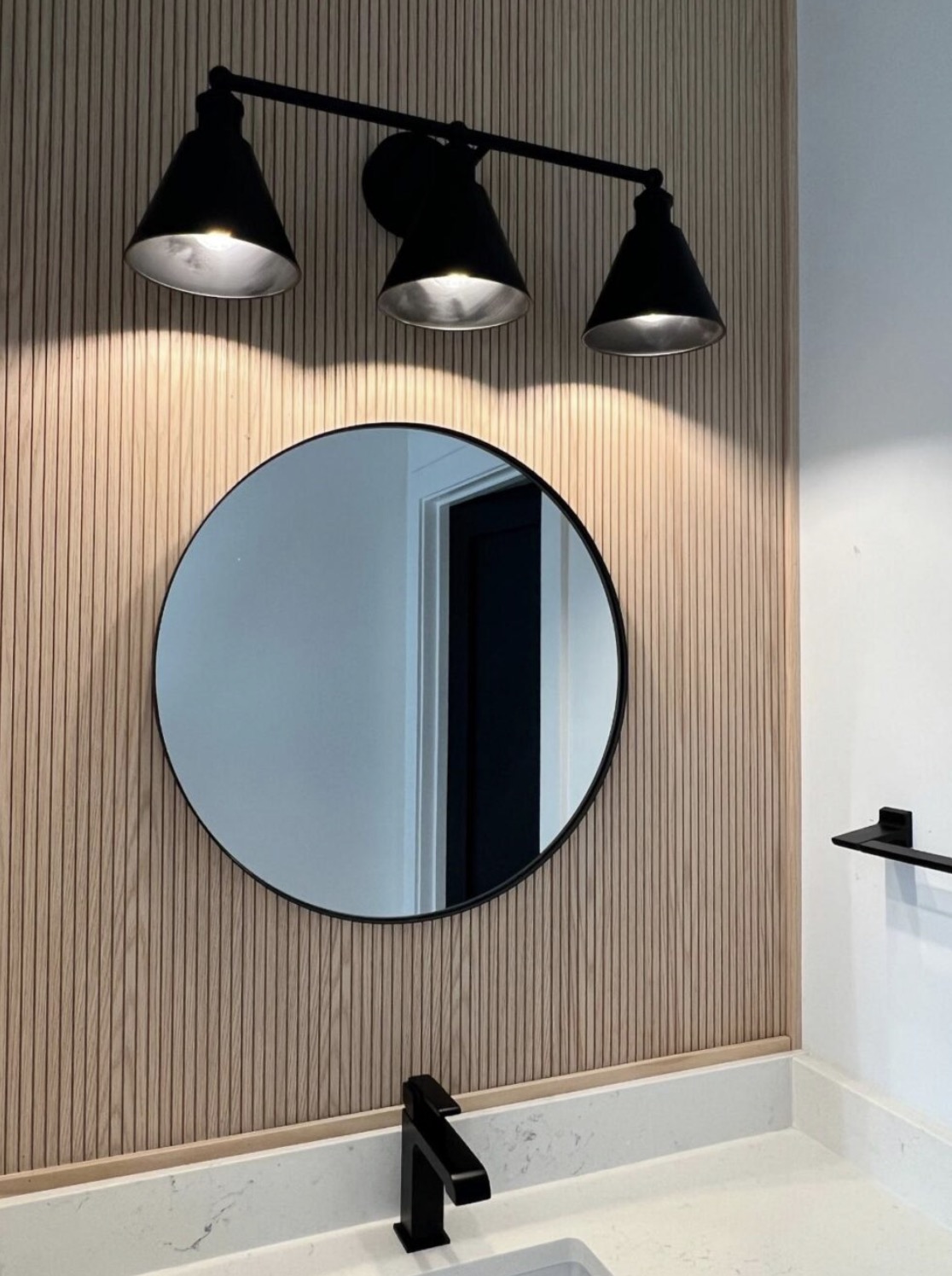 Powder room with fluted white oak wall paneling, round black framed mirror and matte black vanity light