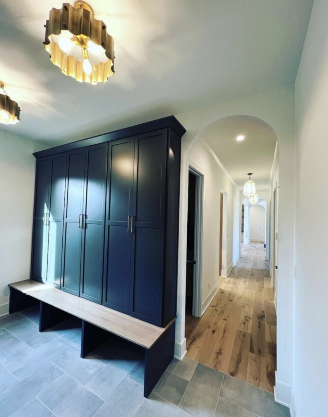 Mudroom with navy custom lockers, oak bench seat and antique brass ceiling fixtures beside an arched hallway