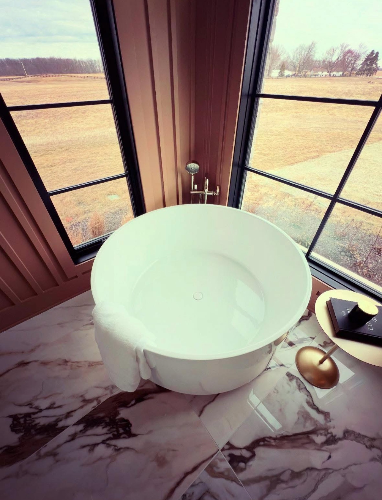 Primary bath with round freestanding soaking tub, marble floors and black framed corner windows over open farmland