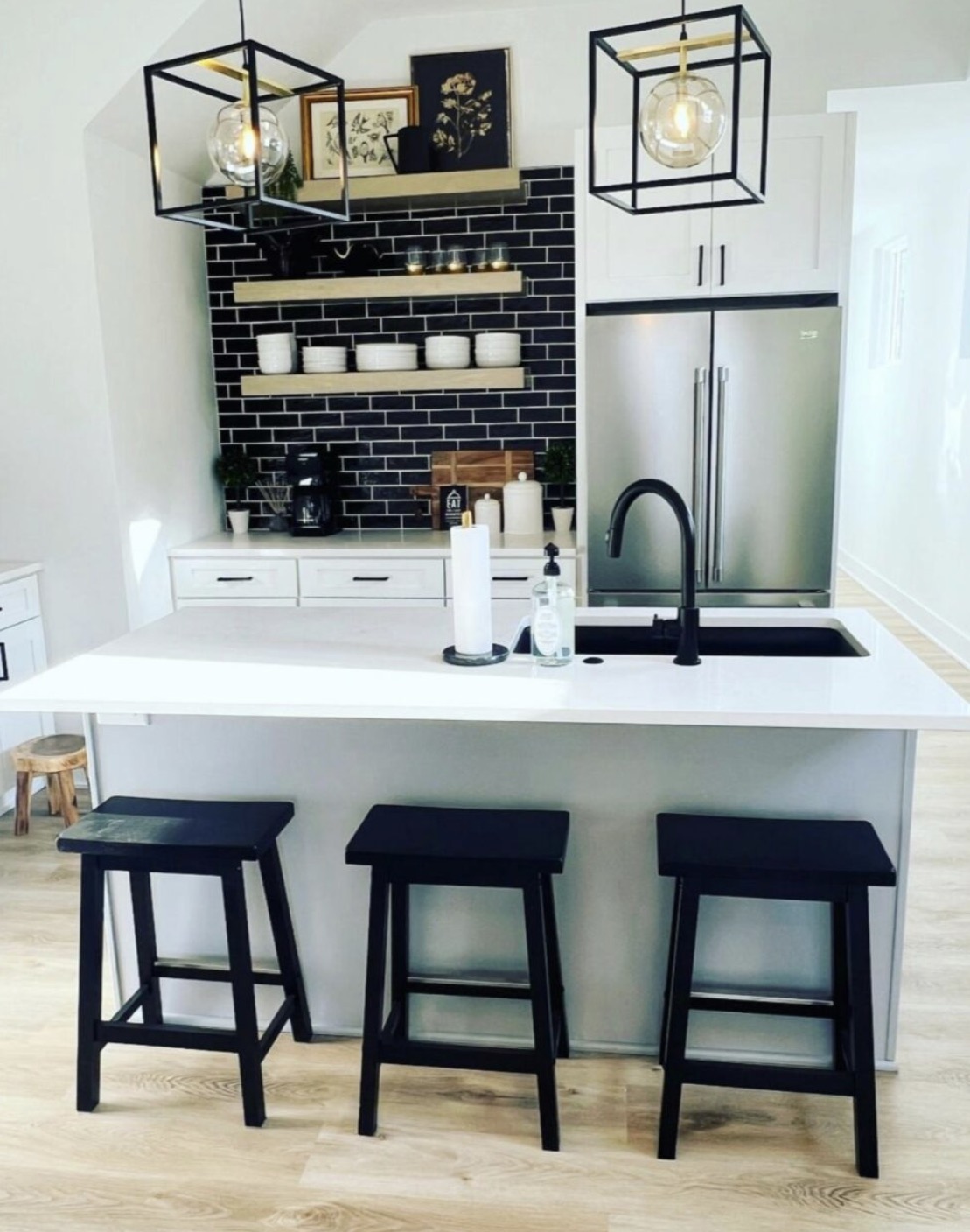 Bright kitchen with quartz island, black subway tile backsplash, floating oak shelves and cage pendant lights